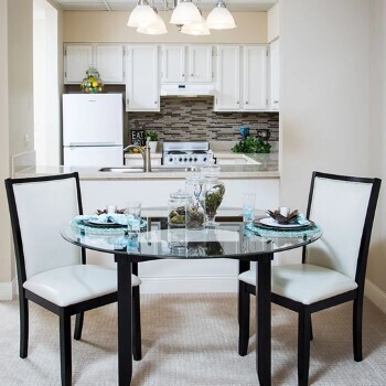 glass dining room table set with dishware. Two chairs are pulled up and the kitchen is visible in the background